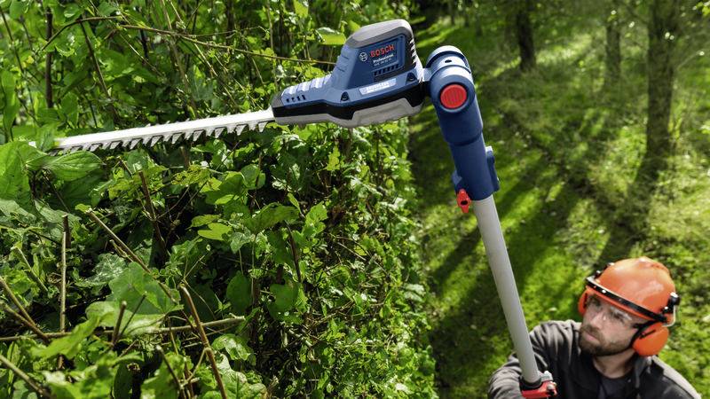 Eine Person verwendet eine Teleskop-Heckenschere in einem Garten, um hohe Hecken zu schneiden. Die Person trägt Schutzkleidung.