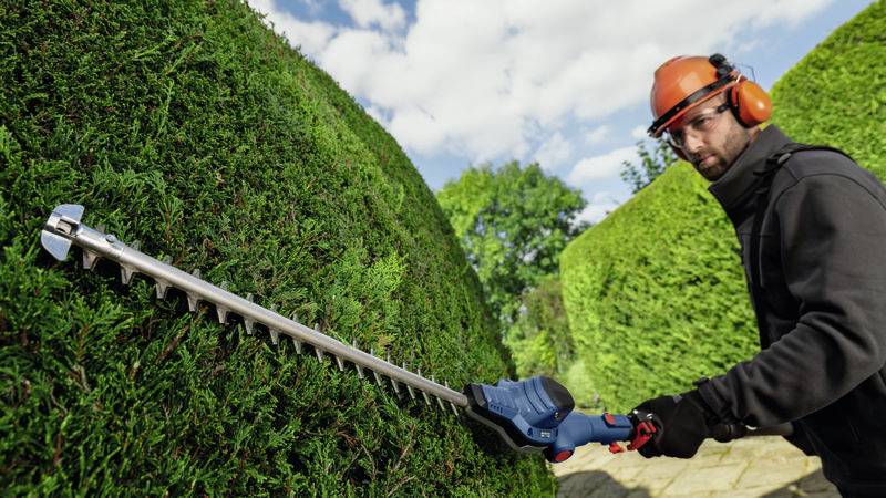 Ein Mann mit Schutzhelm und Gehörschutz schneidet eine Hecke mit einer elektrischen Heckenschere, umgeben von grünen Büschen.