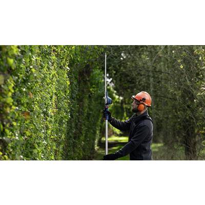 Ein Gärtner mit Schutzhelm und Gehörschutz schneidet eine hohe Hecke mit einer langen Heckenschere in einem Garten.