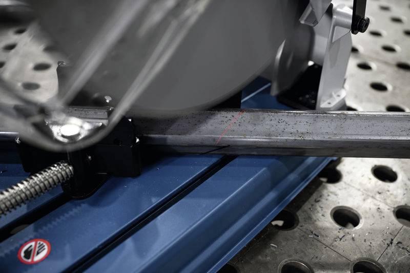 Eine Metallsäge schneidet ein Metallstück auf einem blauen Arbeitstisch. Eine rote Linie zeigt die Schnittführung an.