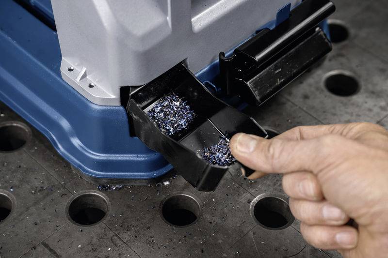Hand entleert Auffangbehälter mit Metallspänen aus einer blauen Maschine auf einem Lochblechtisch.