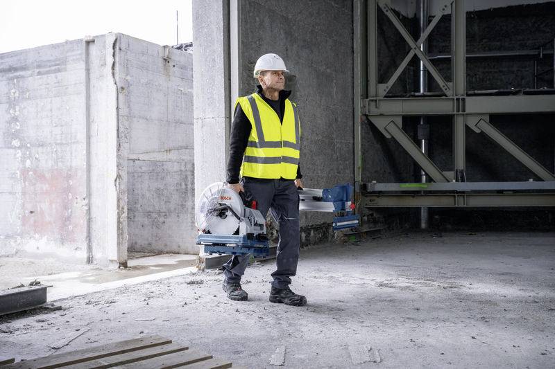 Ein Bauarbeiter in Schutzweste und Helm trägt ein Elektrowerkzeug in einem halbfertigen Gebäude mit Betonwänden.