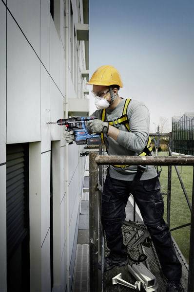Bauarbeiter auf einem Gerüst bohrt mit Akkuschrauber in eine Gebäudeaußenwand. Er trägt Schutzhelm, Brille und Maske.