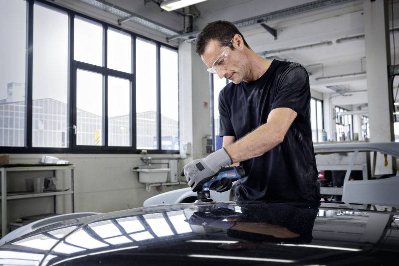 Ein Mann in Schutzbrille und Handschuhen poliert das Dach eines Autos in einer Werkstatt. Im Hintergrund sind große Fenster sichtbar.