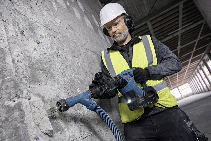 Arbeiter in Warnweste und Helm bedient einen Bohrhammer an einer Betonwand in einem Rohbau.