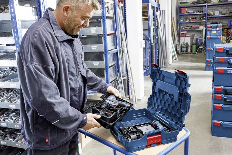 Eine Person öffnet einen blauen Werkzeugkoffer in einer Werkstatt. Der Koffer enthält Elektrowerkzeuge und Zubehör. Regale im Hintergrund.