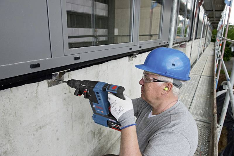 Ein Bauarbeiter mit blauem Schutzhelm und Gehörschutz bohrt mit einer elektrischen Bohrmaschine in eine Betonwand neben einer Fensterfront.