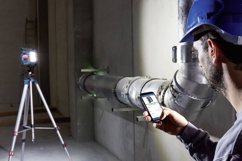 Eine Person in Bauhelm betrachtet ein Smartphone vor einem beleuchteten Metallrohr in einem industriellen Gebäude. Ein Baustrahler erhellt die Szene.