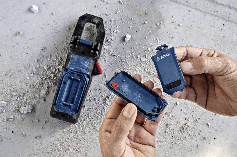 Eine Person hält ein offenes, blaues Bosch-Werkzeug auf einer Baustelle, umgeben von Staub und Schutt.