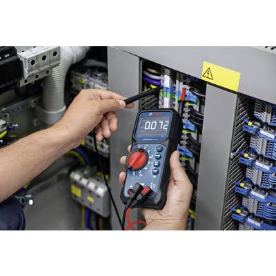 Eine Person misst mit einem digitalen Multimeter in einem Schaltschrank die Spannung. Das Display zeigt '0.72' an.