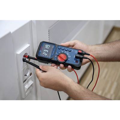 Person misst Spannung mit einem digitalen Multimeter an einer Stromsteckdose. Display zeigt Messwerte.
