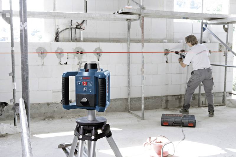 Person arbeitet in einem Raum mit Bauwerkzeug, während im Vordergrund ein Lasergerät auf einem Stativ rote Linien projiziert.