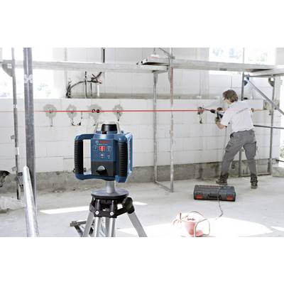 Person arbeitet in einem Raum mit Bauwerkzeug, während im Vordergrund ein Lasergerät auf einem Stativ rote Linien projiziert.
