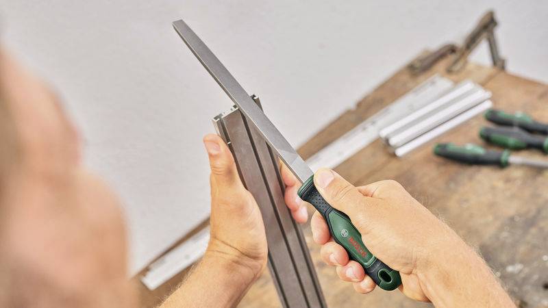 Person feilt mit einer Metallfeile an einem Metallstück. Im Hintergrund liegen Werkzeuge auf einer Werkbank.