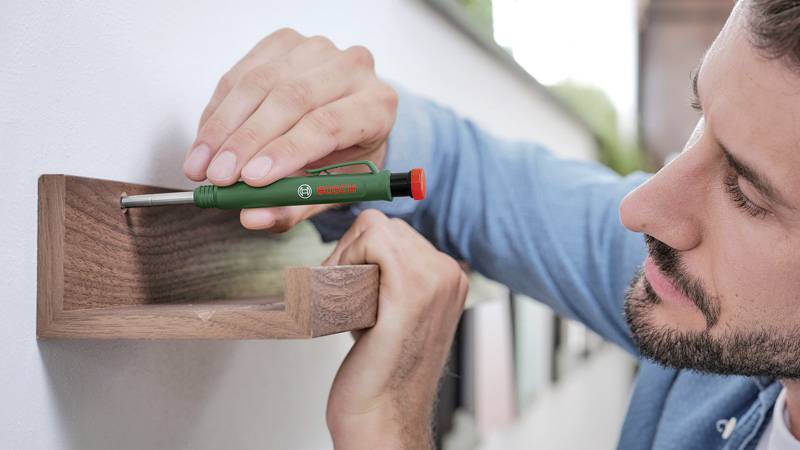 Ein Mann befestigt ein kleines Holzregal an der Wand mit einem Schraubenzieher. Im Hintergrund ist ein unscharfer Außenbereich erkennbar.