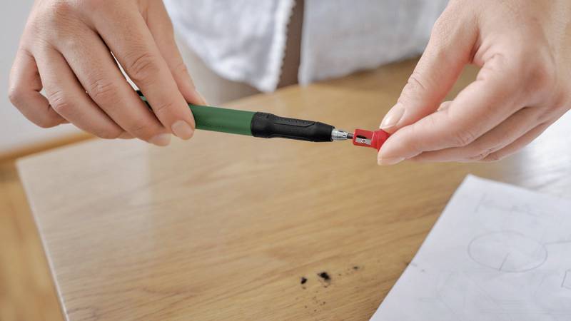 Eine Person steckt eine rote Mine in einen grünen Druckbleistift über einem Holztisch mit Papier und Bleistiftspuren.