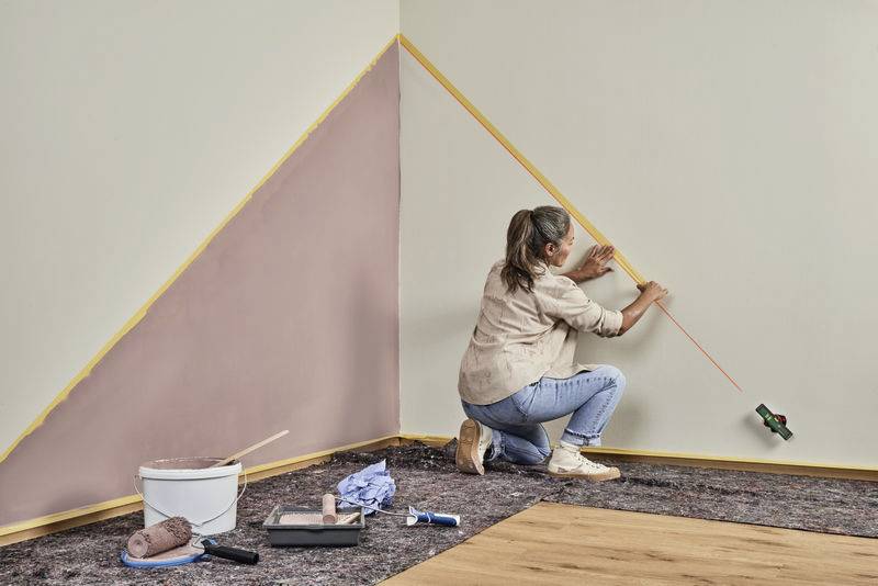 Eine Frau streicht eine Wand in einem Raum. Sie verwendet ein langes Maßband, um eine Diagonale mit rosa Farbe abzukleben.