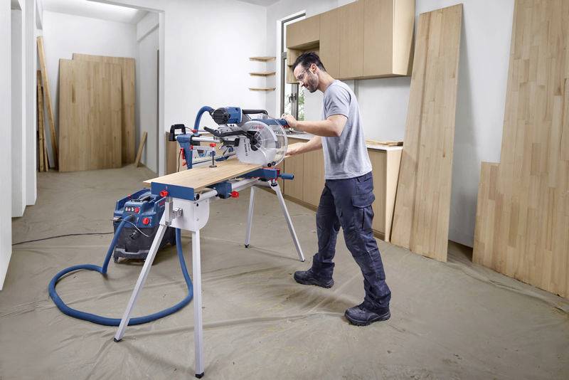 Ein Mann bedient eine Kreissäge in einer Werkstatt, um ein Holzbrett zu schneiden. Im Hintergrund sind Holzplatten und Schränke sichtbar.