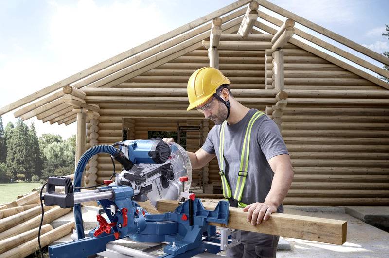 Ein Bauarbeiter mit Helm und Schutzbrille schneidet ein Holzbrett mit einer elektrischen Kappsäge vor einem Holzhaus.