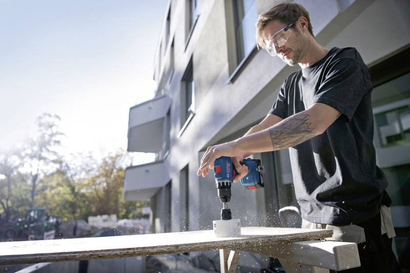Ein Mann bohrt mit einer elektrischen Bohrmaschine ein Loch in ein Holzbrett vor einem modernen Wohngebäude.