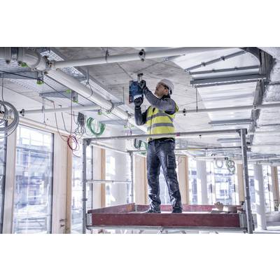 Ein Bauarbeiter in Sicherheitsweste und Helm steht auf einem Gerüst und bohrt Löcher in die Decke eines halbfertigen Gebäudes.
