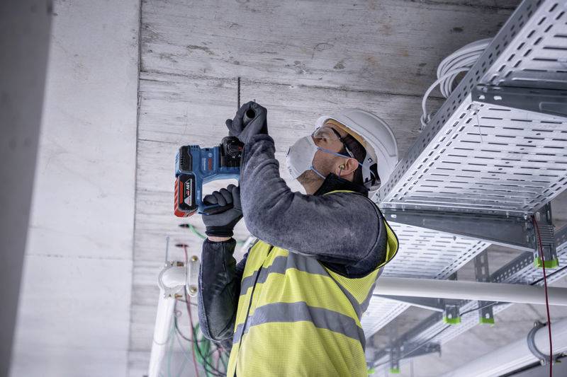 Ein Bauarbeiter mit Schutzhelm und Weste bohrt in eine Decke. Kabel und Rohre sind sichtbar. Trägt Maske und Arbeitshandschuhe.