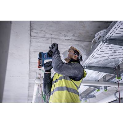 Ein Bauarbeiter mit Schutzhelm und Weste bohrt in eine Decke. Kabel und Rohre sind sichtbar. Trägt Maske und Arbeitshandschuhe.