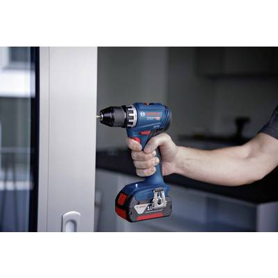 Eine Hand hält einen blauen Akku-Bohrschrauber vor einem Fenster, bereit zur Benutzung. Im Hintergrund ist ein unscharfes Zimmer zu sehen.