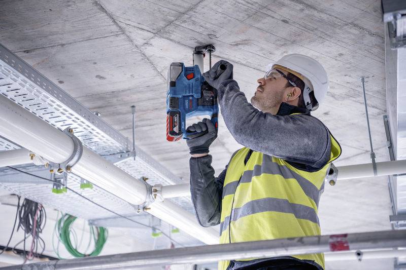 Arbeiter mit Schutzhelm und Warnweste bohrt mit einer Elektrowerkzeugmaschine in die Decke, um Kabel zu installieren.