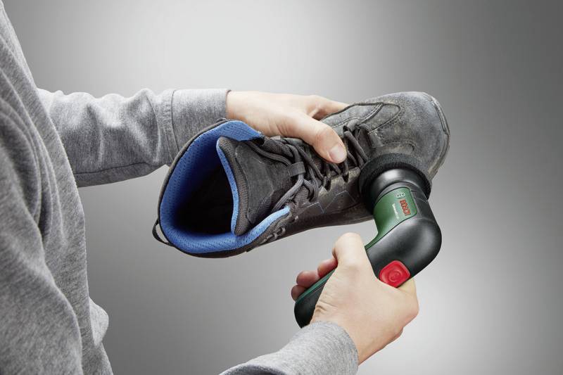 Person hält einen Schuh in der Hand und reinigt ihn mit einem elektrischen Gerät. Der Hintergrund ist neutral.