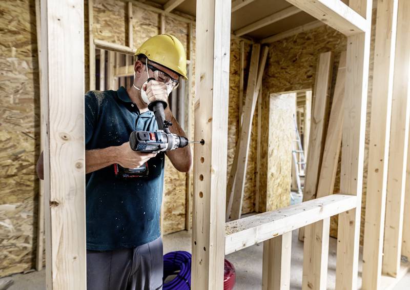 Ein Arbeiter mit Schutzhelm und Maske benutzt eine Bohrmaschine, um Löcher in einen Holzrahmen zu bohren, in einem Rohbau-Gebäude.