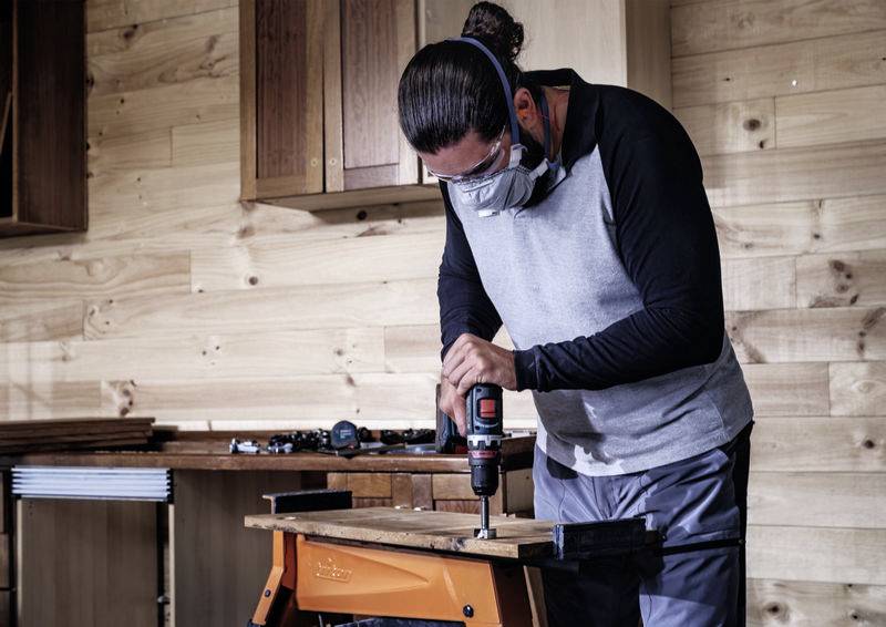 Mann mit Schutzbrille und Maske benutzt eine Bohrmaschine, um ein Loch in ein Holzbrett zu bohren, in einer Werkstatt mit Holzwänden.