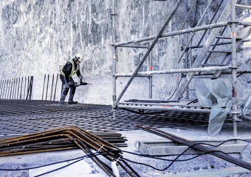 'Bauarbeiter in Schutzkleidung arbeitet an Stahlbewehrung neben einem Gerüst in einer Baustelle, Betonwand im Hintergrund.'