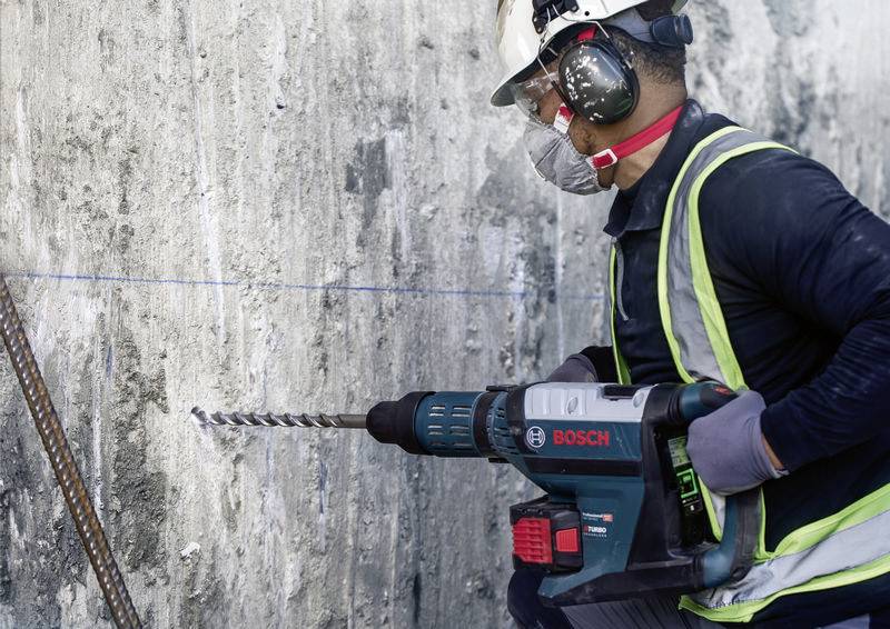 Ein Bauarbeiter mit Schutzkleidung und Gehörschutz verwendet eine Bosch-Bohrmaschine, um in eine Betonwand zu bohren.