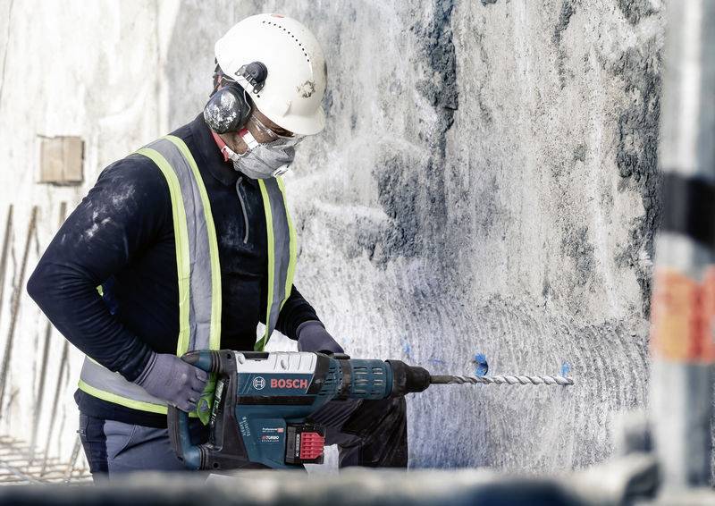 Ein Bauarbeiter bohrt mit einer elektrischen Bohrmaschine in eine Betonwand. Der Arbeiter trägt Schutzkleidung und einen Helm.