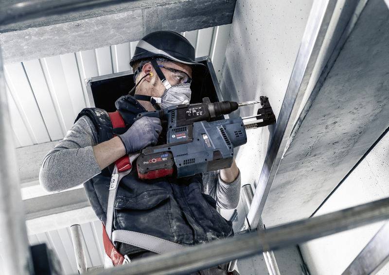 Ein Bauarbeiter mit Schutzhelm und Maske bohrt mit einem Elektrowerkzeug in eine Betonwand.