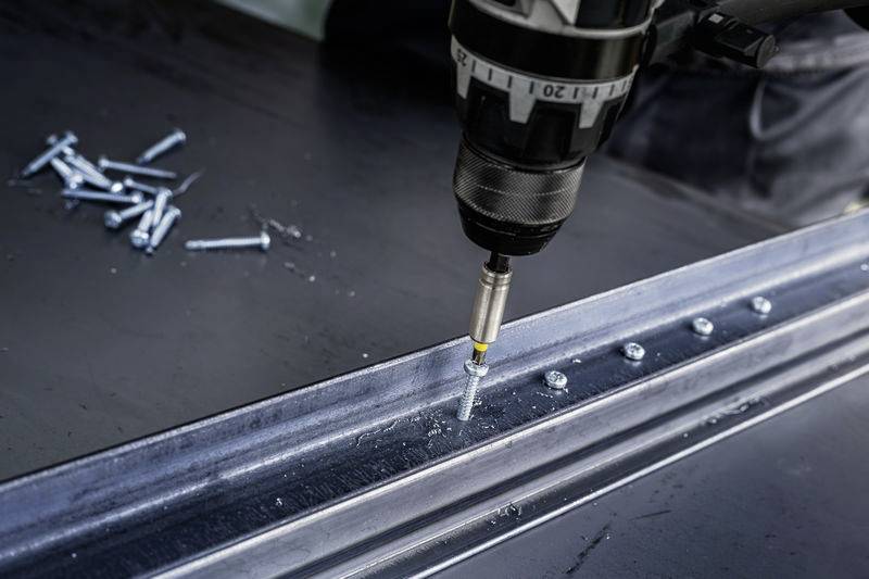 Eine Bohrmaschine zieht Schrauben in ein Metallblech ein, Schrauben liegen daneben. Es veranschaulicht den Montageprozess.