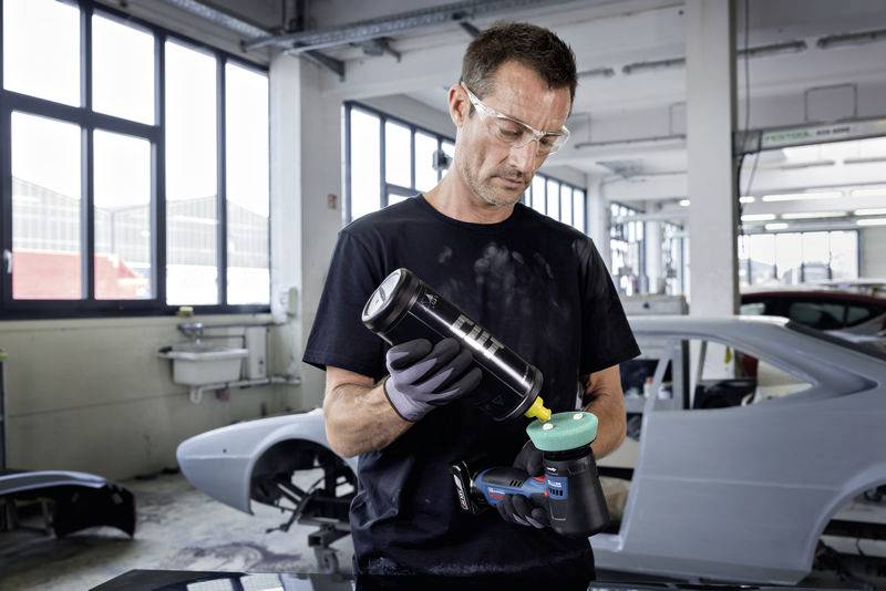 Ein Mann in einer Autowerkstatt trägt Schutzbrille und Handschuhe, während er ein Werkzeug mit Polierscheibe vorbereitet. Auf einem Werkstattboden stehen Autos im Hintergrund.