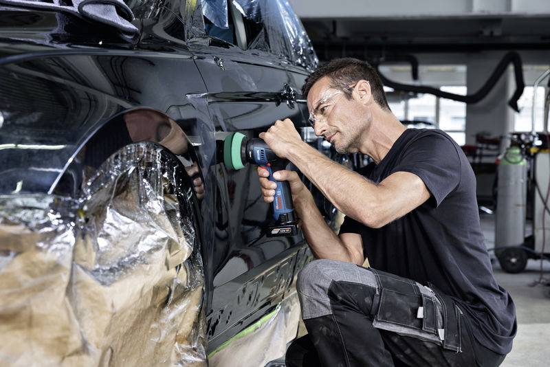 Ein Mann poliert ein Auto mit einer Maschine in einer Werkstatt. Er trägt Schutzbrille und schwarze Kleidung. Das Auto ist teilweise abgedeckt.