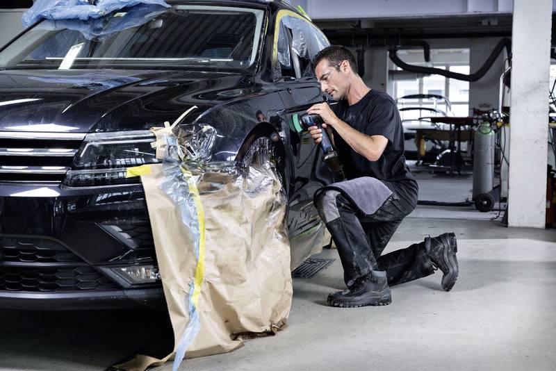 Ein Mechaniker arbeitet in einer Werkstatt an der vorderen Stoßstange eines schwarzen Autos. Der Bereich ist mit Folie und Papier abgedeckt.