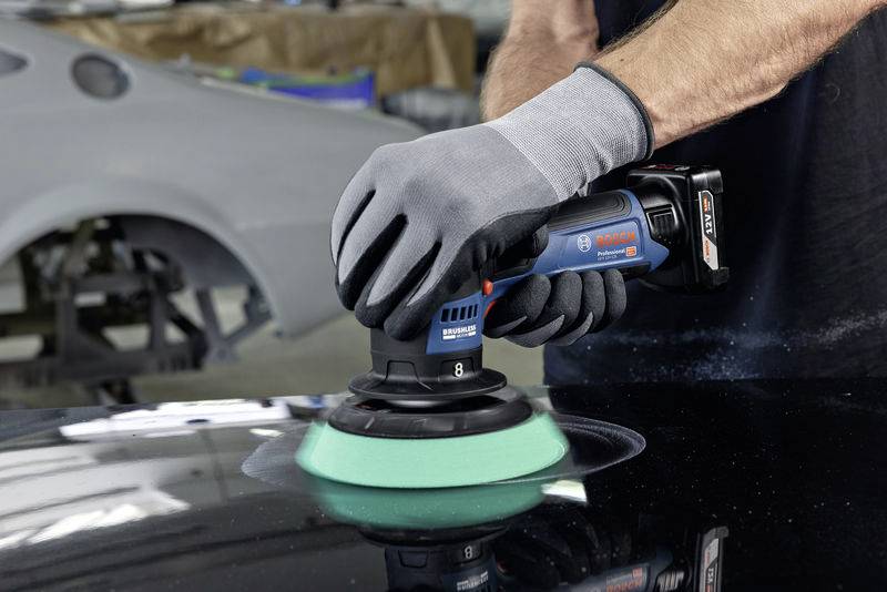 Eine Person poliert eine Autokarosserie mit einer kabellosen Poliermaschine. Im Hintergrund ist ein weiteres Fahrzeug sichtbar.