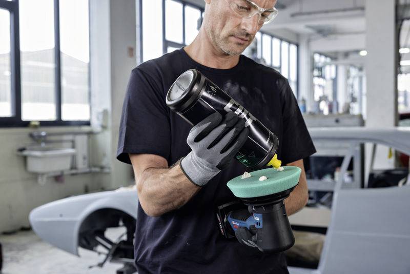 Ein Mann in einer Werkstatt trägt Schutzbrille und Handschuhe und bereitet eine Poliermaschine mit Polierpaste vor; ein Auto im Hintergrund.