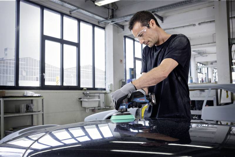 Ein Mann mit Brille und Handschuhen poliert die Oberfläche eines Autos in einer Werkstatt. Er verwendet eine elektrische Poliermaschine.