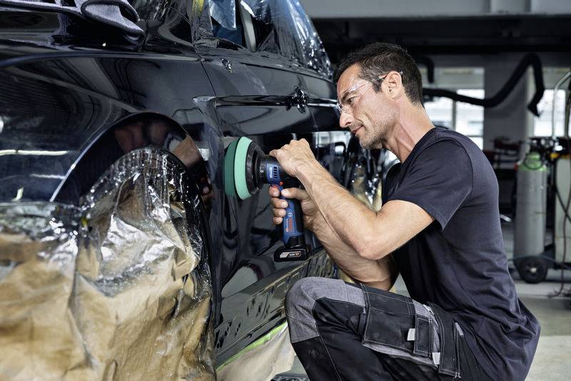 Ein Mann poliert mit einer Poliermaschine die schwarze Karosserie eines Autos in einer Werkstatt.
