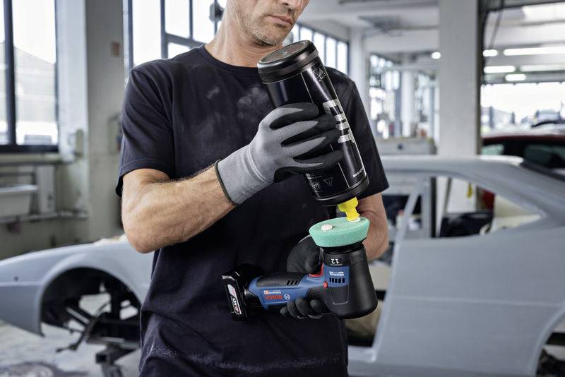 Eine Person in Arbeitskleidung benutzt eine elektrische Poliermaschine in einer Autowerkstatt, während ein Auto im Hintergrund sichtbar ist.