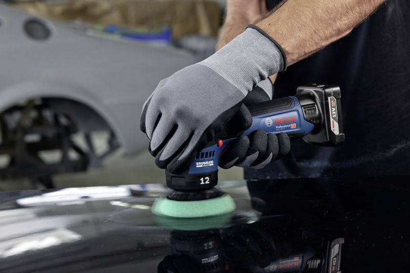 Person poliert einen Autolack mit einer elektrischen Poliermaschine in einer Werkstatt. Hände tragen graue Handschuhe.