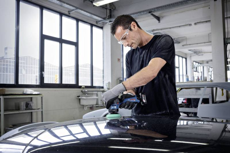 Ein Mann poliert mit einer Maschine die Motorhaube eines Autos in einer Werkstatt. Er trägt Schutzbrille und Handschuhe.