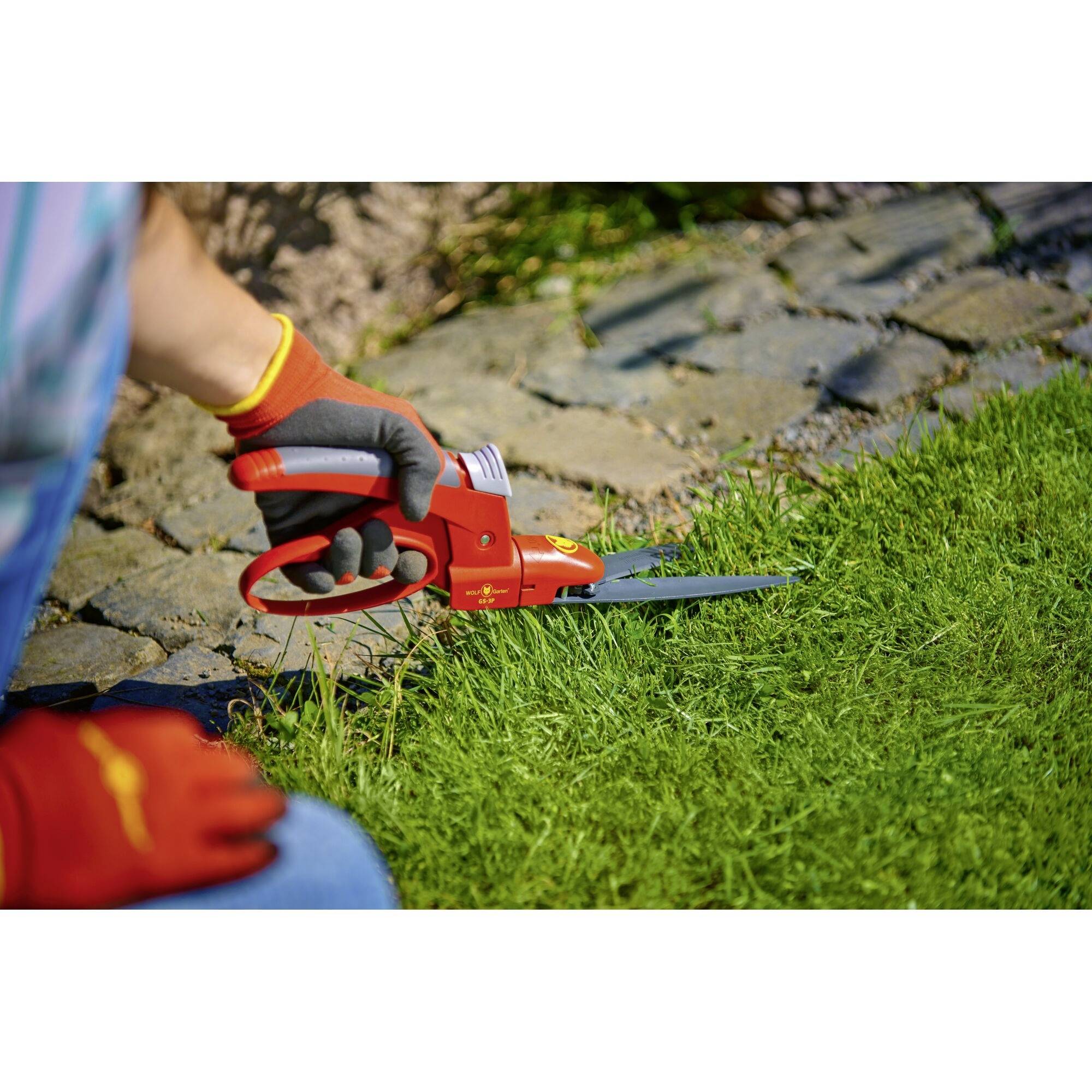 Person schneidet mit roten Handscheren Gras neben einem Steinweg. Sie trägt rote Gartenhandschuhe und sitzt auf dem Boden.