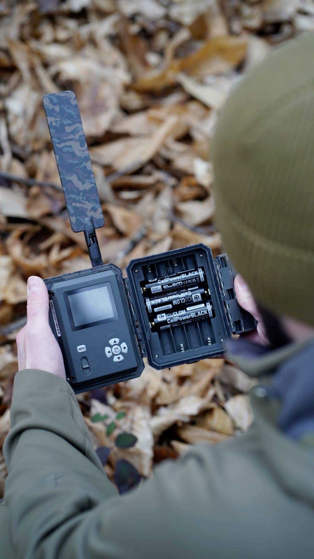 Eine Person hält ein geöffnetes Wildkamera-Gerät mit Batteriefach und Bildschirm, umgeben von herbstlichen Blättern im Wald.