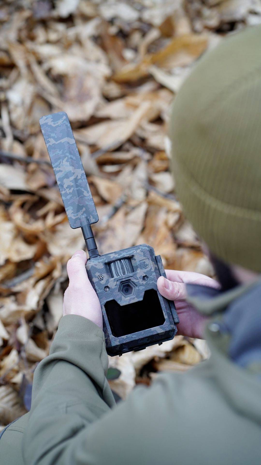 Eine Person hält eine getarnte Wildkamera im herbstlichen Laub. Sie trägt eine grüne Jacke und eine Mütze. Die Kamera hat eine lange Antenne.